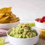 A white bowl piled with green guacamole with small bits of chopped tomatoes.