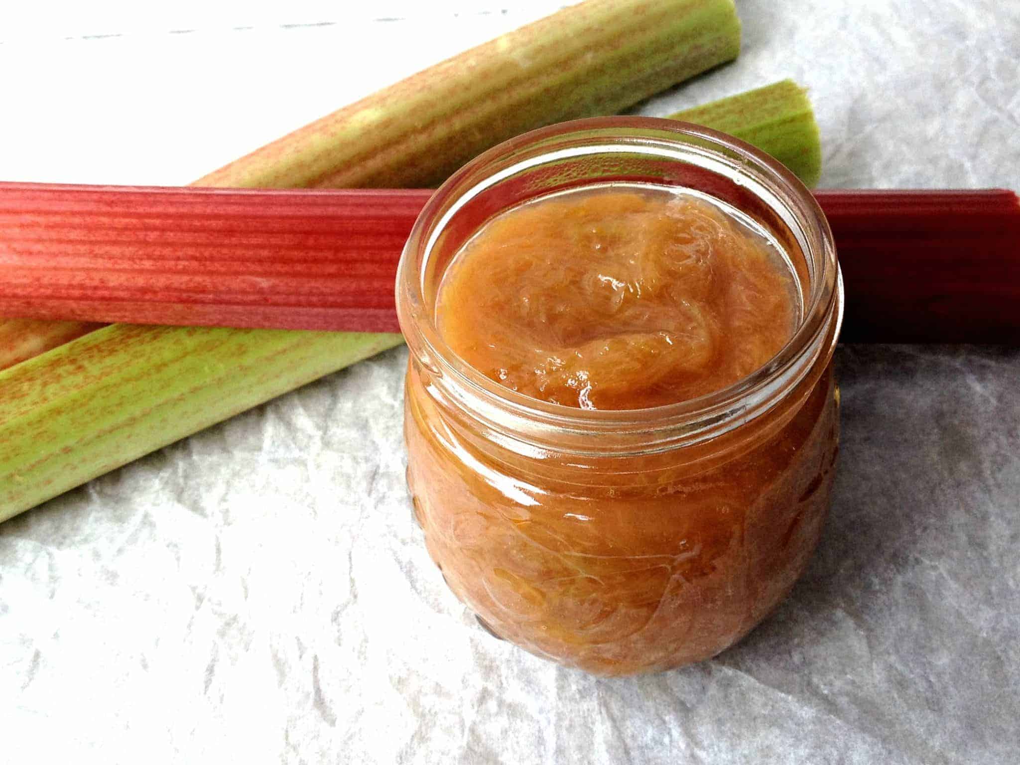 A jar full of cooked rhubarb with fresh stalks of rhubarb next to jar.