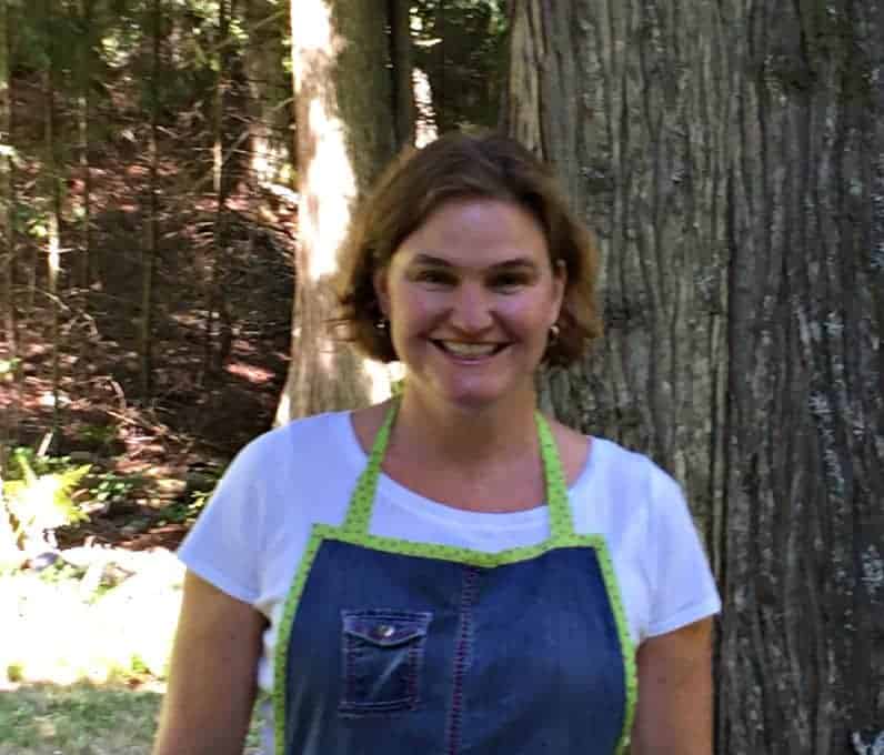 Kathy wearing an apron and standing in front of a tree.