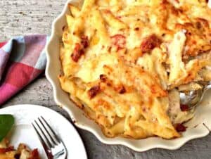Baked cheesy penne pasta with chicken and roasted tomatoes in a white baking dish.