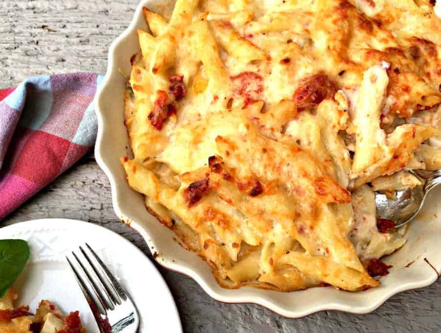 Baked cheesy penne pasta with chicken and roasted tomatoes in a white casserole dish.