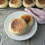 A plate with two english muffins with a baking rack filled of muffins behind the plate.