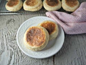 A plate with two english muffins with a baking rack filled of muffins behind the plate.
