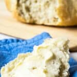 A piece of rustic bread slathered with butter with a round loaf of bread in the background.