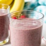 A glass filled with a smoothie with bananas and fruit behind the glass.