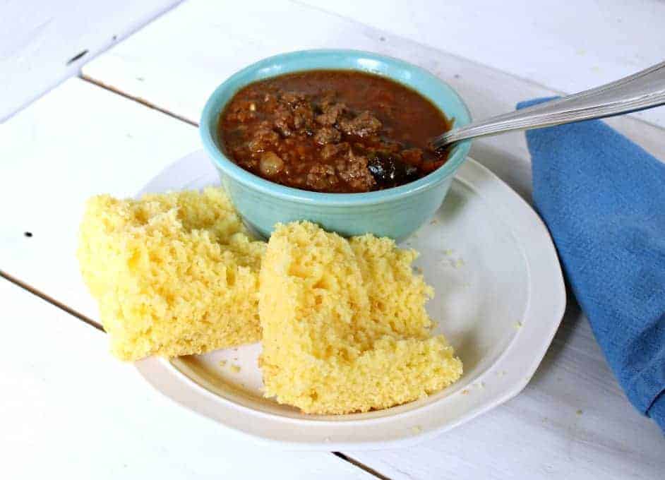 A piece of corn bread cut in half on a plate next to a small bowl of chili.