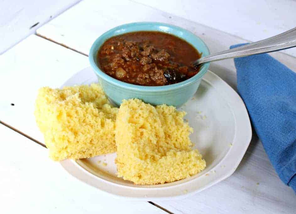 A piece of corn bread cut in half on a plate next to a small bowl of chili.