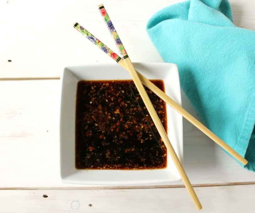 A square white bowl with a dark sauce with two chopsticks crossed over the top of the bowl.