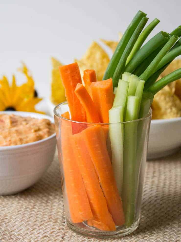 A glass filled with freshly cut carrots, celery and green onions next to a bowl of hummus. 
