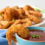 Golden coconut shrimp hanging off the edge of a small bowl filled with a red sauce.