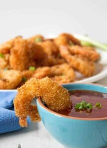 Golden coconut shrimp hanging off the edge of a small bowl filled with a red sauce.