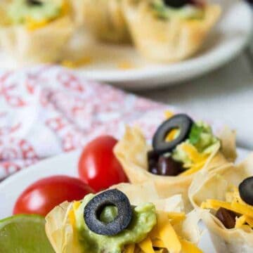 Small pastry cups filled with guacamole, cheese, tomatoes and a sliced black olive.