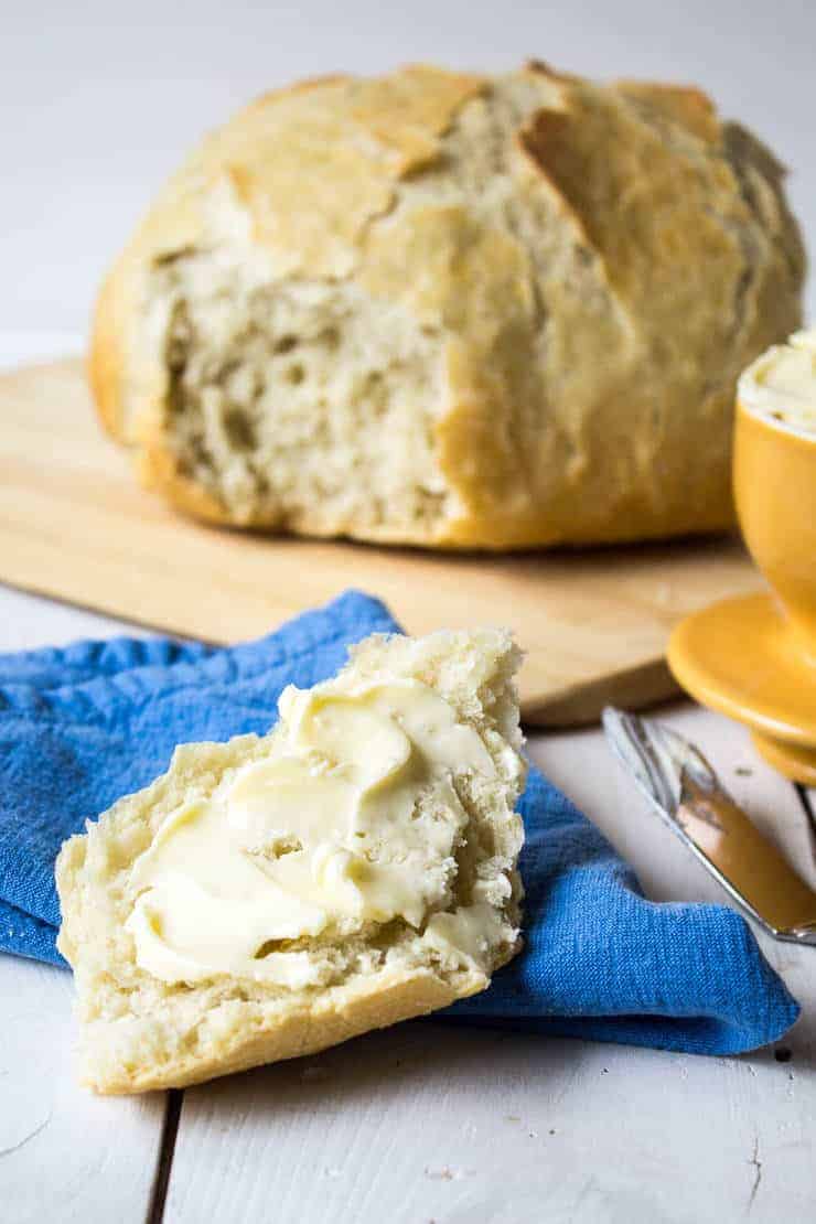 A piece of rustic bread spread with butter with a round loaf of the bread in the background.