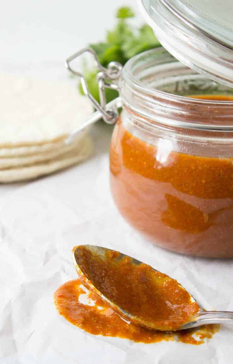 A spoon covered with a red sauce and a glass jar next to the spoon.