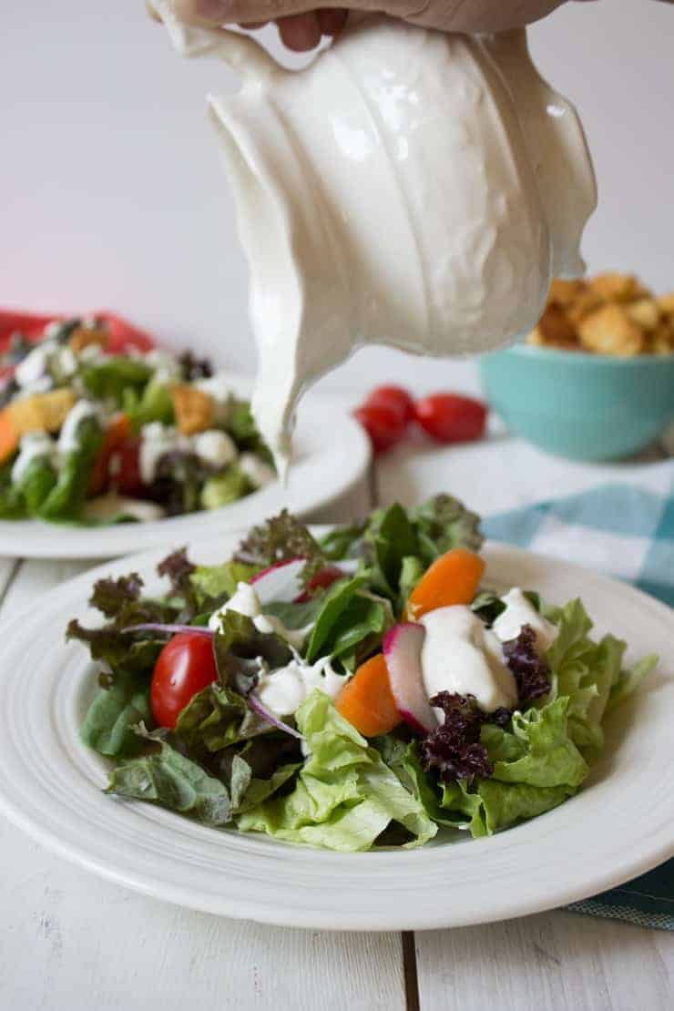 Salad dressing being poured out of a small white pitcher onto a green salad.