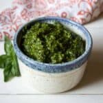 A small bowl filled with a dark green pesto with a basil leaf next to the bowl.