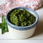 A small bowl filled with a dark green pesto with a basil leaf next to the bowl.