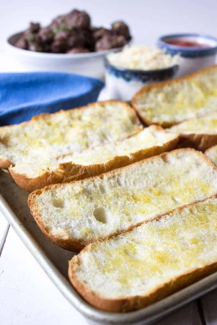 Buttered hoagie rolls on a baking sheet.