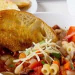 A bowl of soup filled with pasta, beans and vegetables served with a piece of toasted bread.