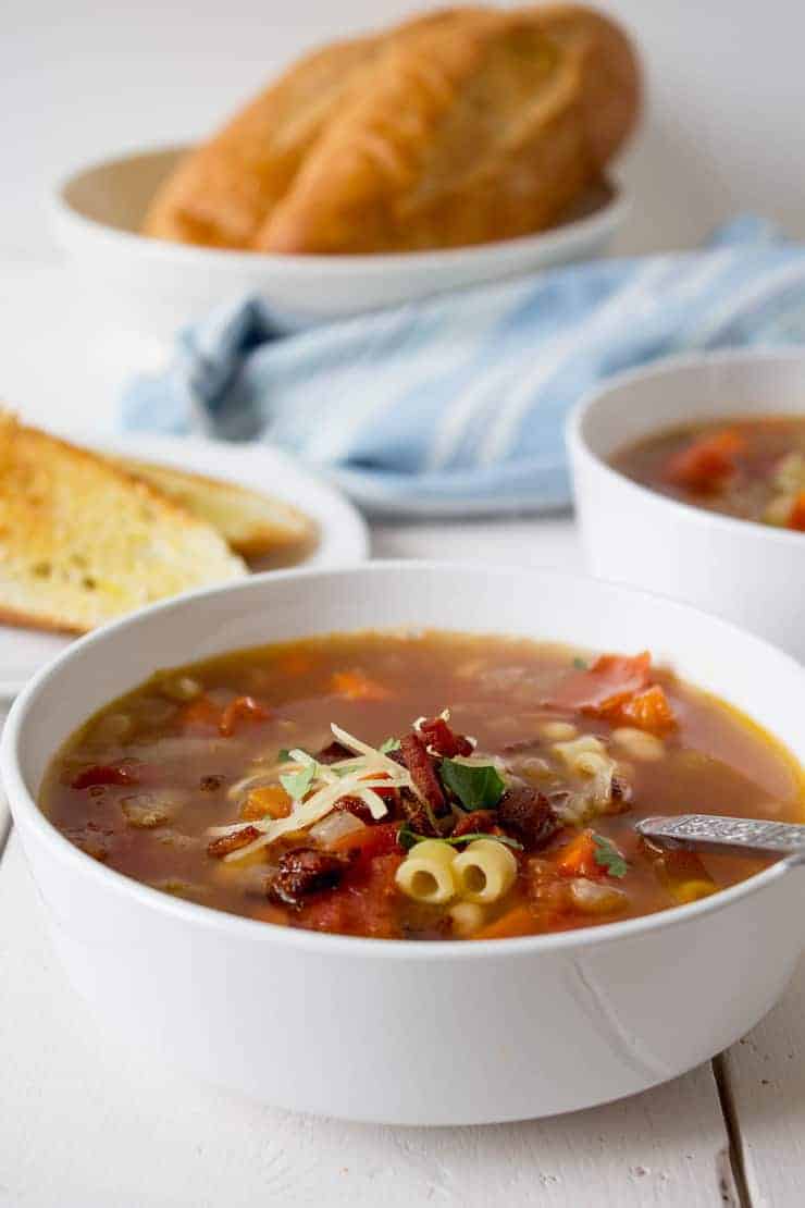 Soup bowl filled with a broth, pasta and beans and toasted bread on a plate next to soup.