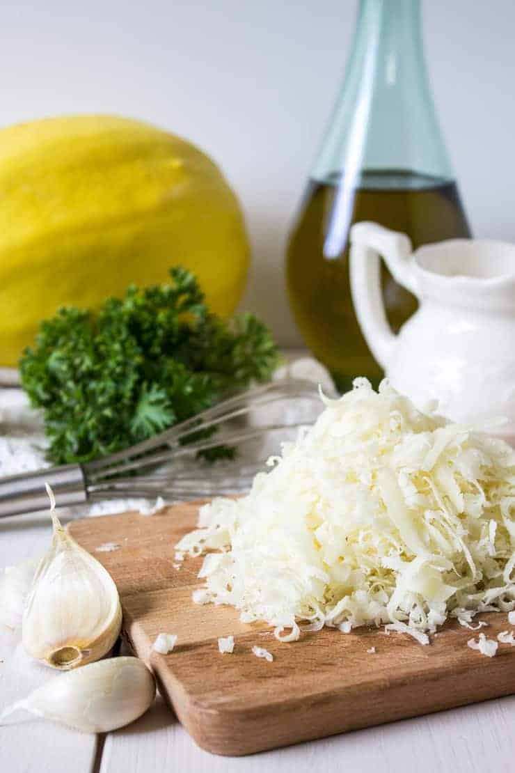 Grated Parmesan cheese, garlic cloves, fresh parsley and a spaghetti squash on a white board.