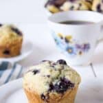 A muffin with blueberries on a small white plate with a teacup in the background.