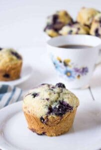 A muffin with blueberries on a small white plate with a teacup in the background.