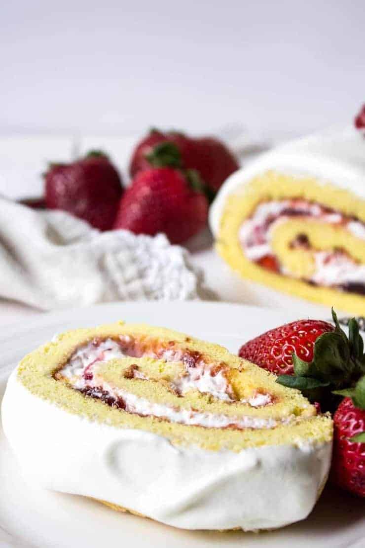 A slice of strawberry swiss roll cake served on a white plate along with fresh strawberries. 