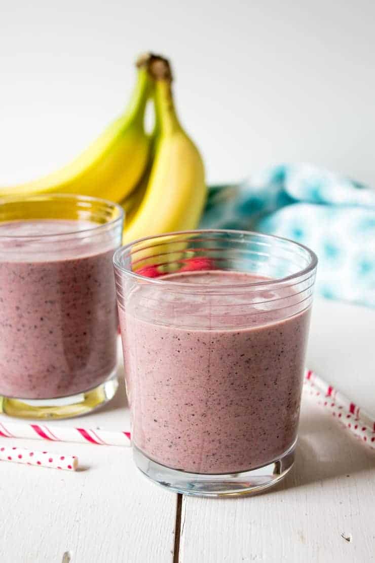 Two glasses filled with a red colored smoothie and fresh bananas in the background. 