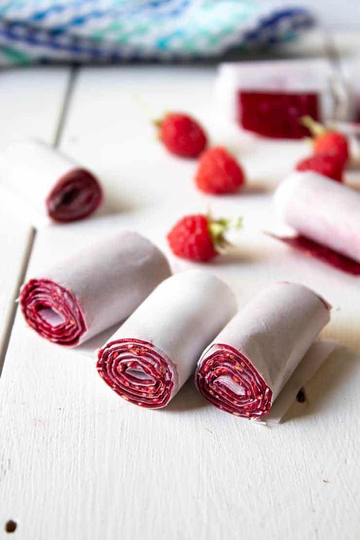 Raspberry Fruit roll ups on a white board with fresh raspberries behind them.