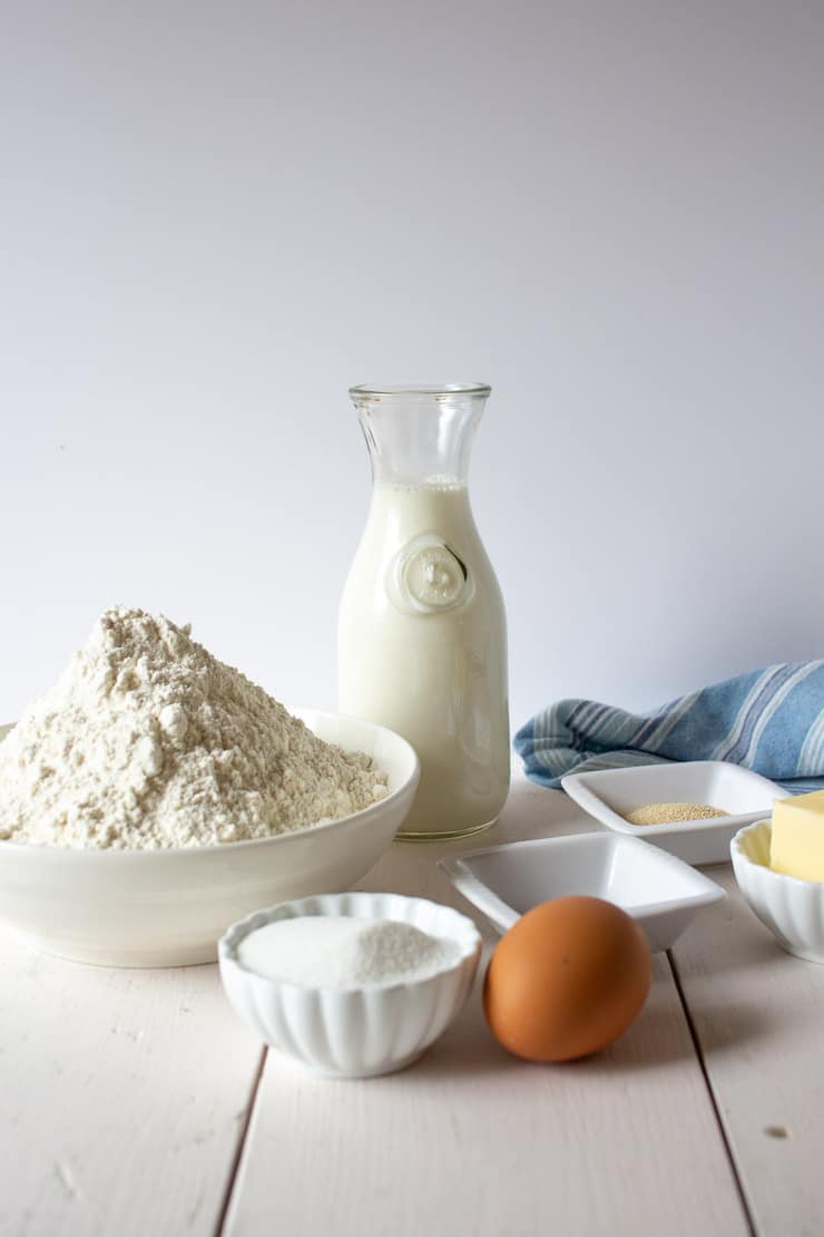 Milk, flour, sugar, egg, yeast, butter and salt on a white board.