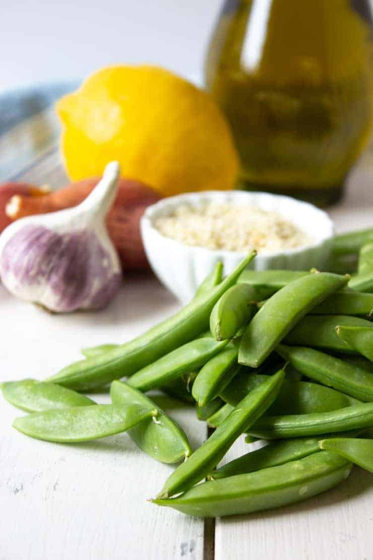 sugar snap peas, garlic, shallots, lemon, olive oil and sesame seeds on a white board.