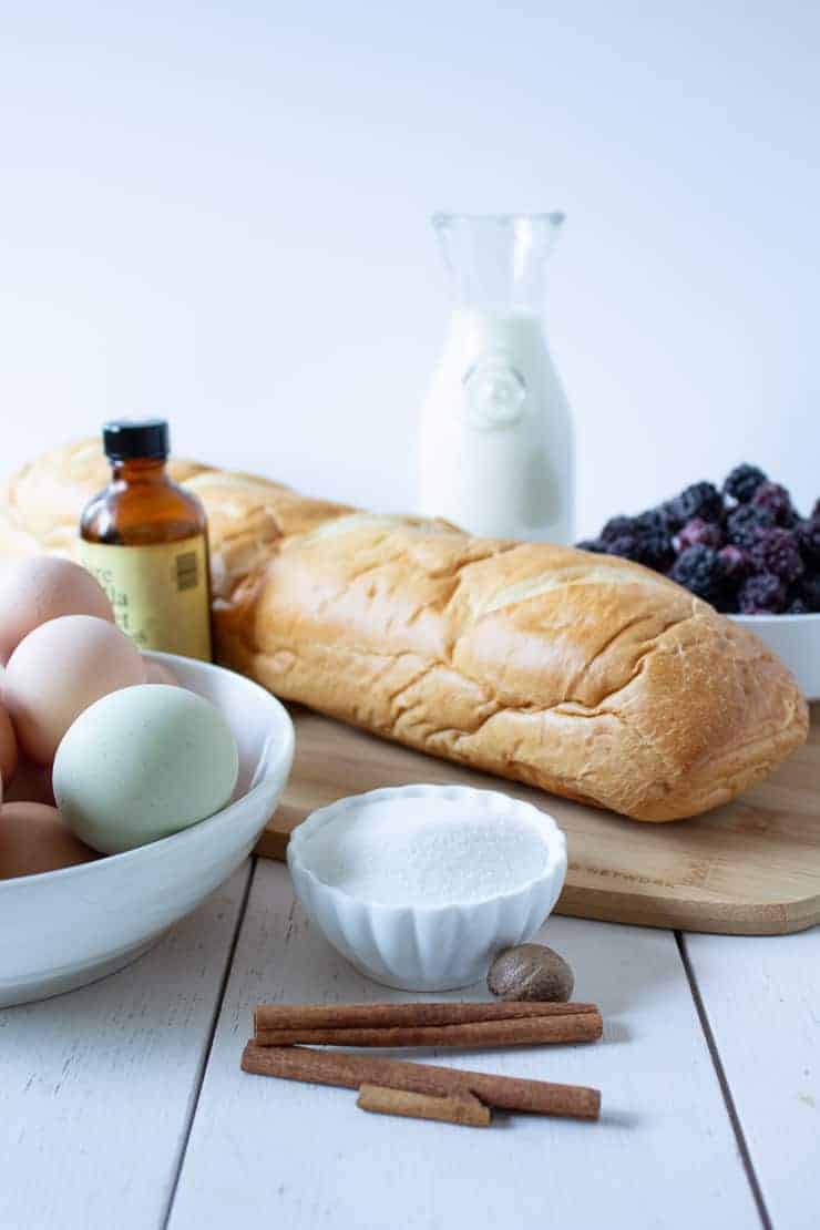 A loaf of french bread next to a bowl of eggs, sugar, cinnamon sticks and a bottle of vanilla.