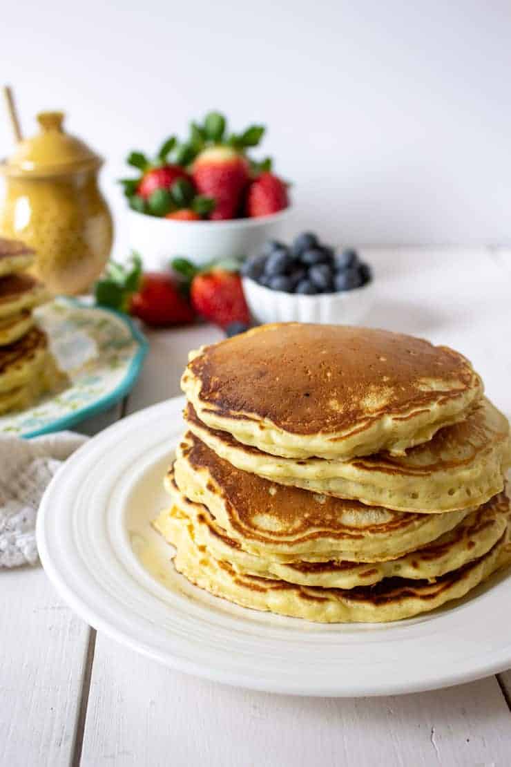 A stack of pancakes on a white plate with fresh berries in the background.