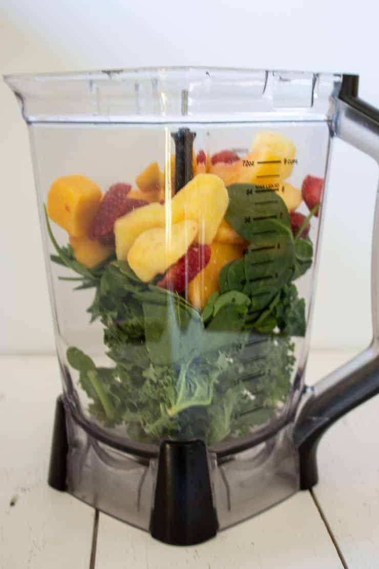 A blender full of frozen fruit, fresh spinach, and fresh kale for making a green smoothie.