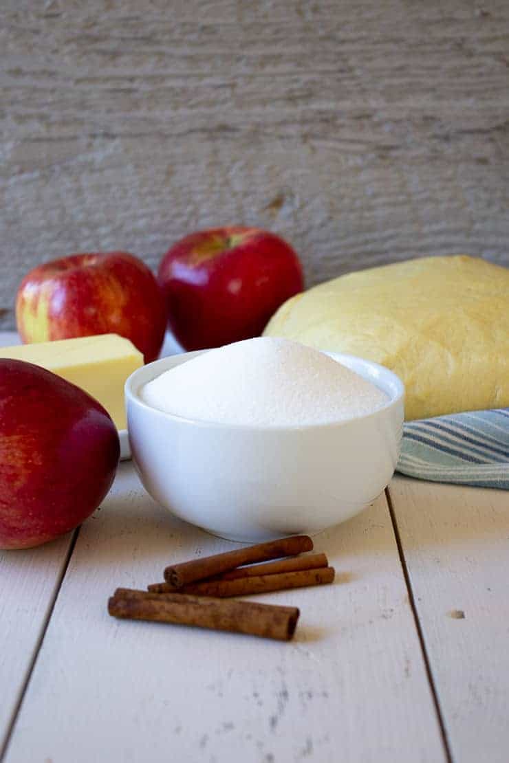 Red apples next to a stick of butter, a bowl of sugar and three cinnamon sticks.