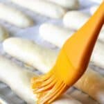 Long sticks of bread dough on a baking sheet with an orange brush adding butter to the tops.