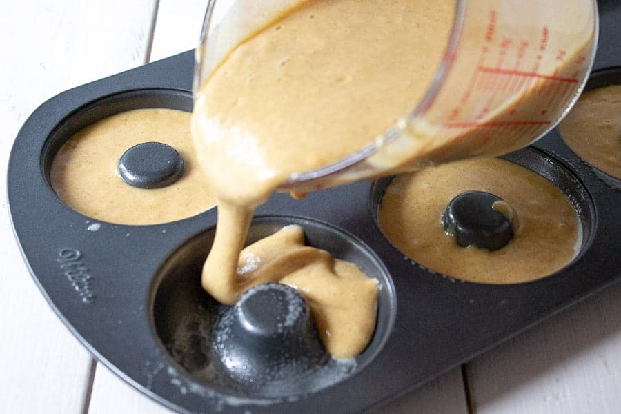 A donut pan being filled with batter poured from a glass measuring cup.