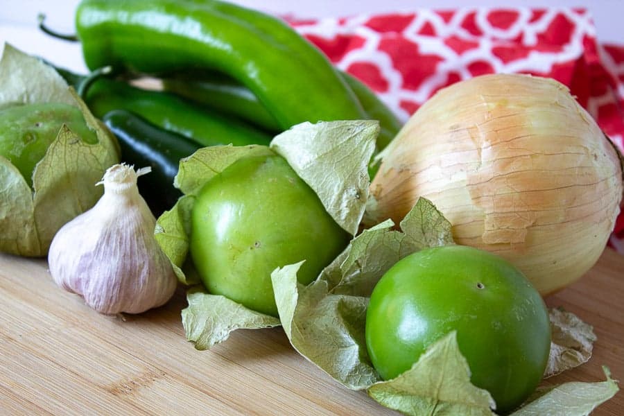 Anaheim peppers, tomatillos, onion and garlic arranged on a wooden cutting board. 