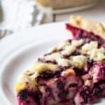 A slice of berry pie on a white plate with a whole pie behind the slice.