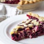 Slice of berry pie on a white plate with the whole pie behind the slice of pie.