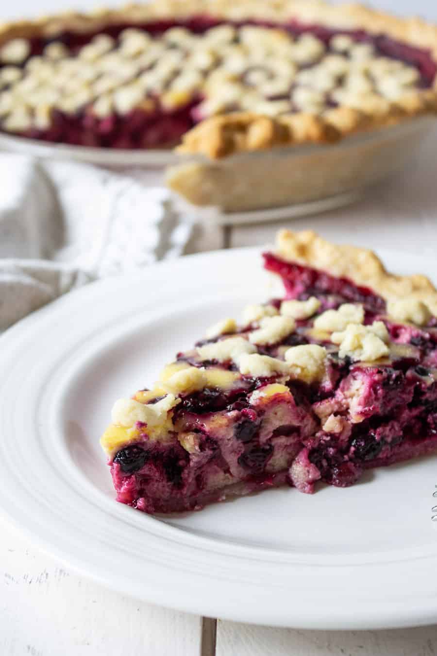 Slice of berry pie on a white plate with the whole pie behind the slice of pie.