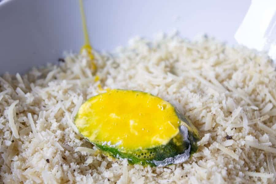 Zucchini slice covered with egg in a bowl with a breading mixture. 