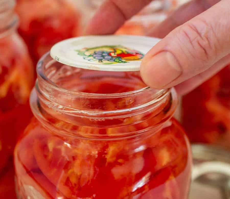 A canning lid being placed on top of a jar of tomatoes.