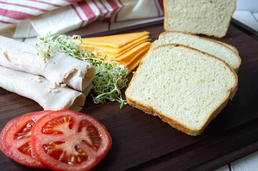 A wooden board topped with sliced bread, tomatoes, turkey, cheese and sprouts. 