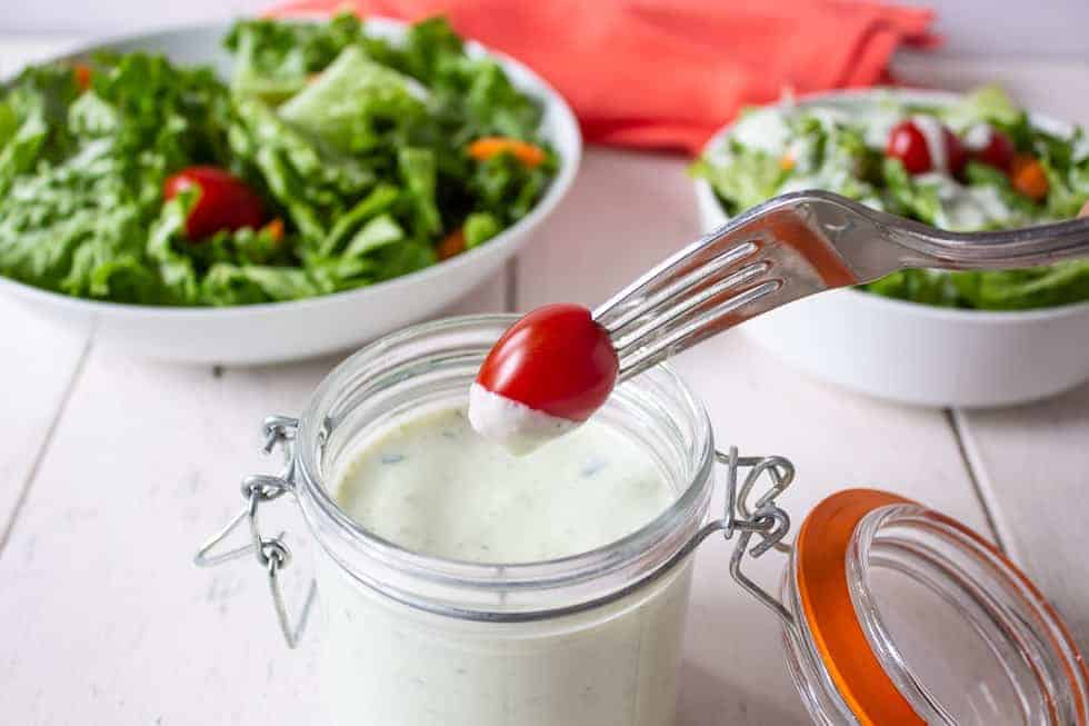 A small red tomato on the end of a fork being dipped into a salad dressing. 