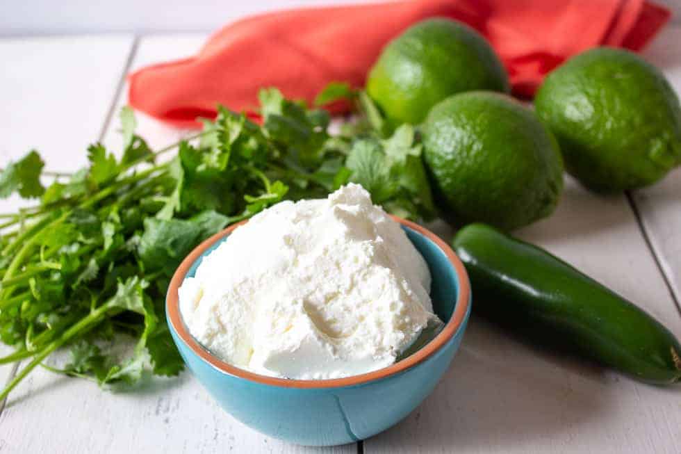 A bowl filled with greek yogurt with fresh cilantro, limes and a jalapeno pepper surrounding the bowl. 