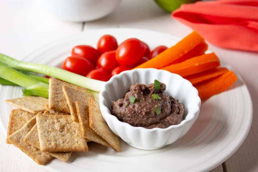 A small dish with a dark colored dip and fresh veggies and crackers on a plate. 