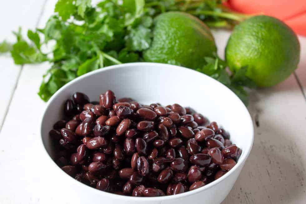 Black beans in a bowl with fresh cilantro and limes behind the bowl. 