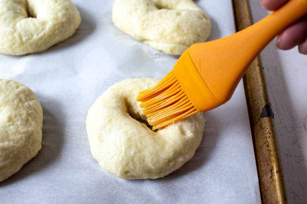 An orange pastry brush adding an egg wash to an uncooked bagel. 